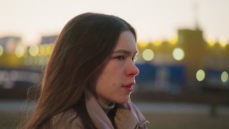 side shot of a young woman wearing a peach jacket, with her mouth slightly open, lost in thought. the soft evening light and blurred urban background add to the contemplative mood