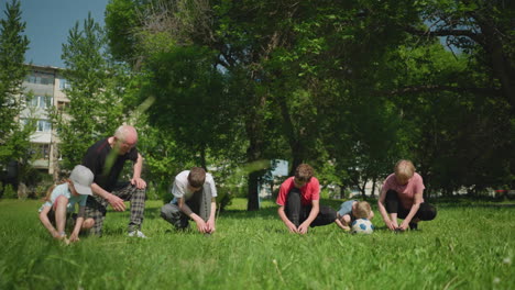 grandparents and their grandsons are tying shoelaces on a grassy field, the youngest boy rests his head on a soccer ball while the grandfather transitions to kneeling on one knee