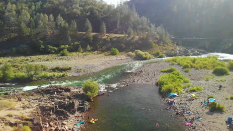 Flying-over-a-swimming-hole-in-the-American-River-in-Auburn,-California---surrounded-by-green-trees,-mountains-and-filled-with-people-swimming-and-enjoying-their-summer-weekend