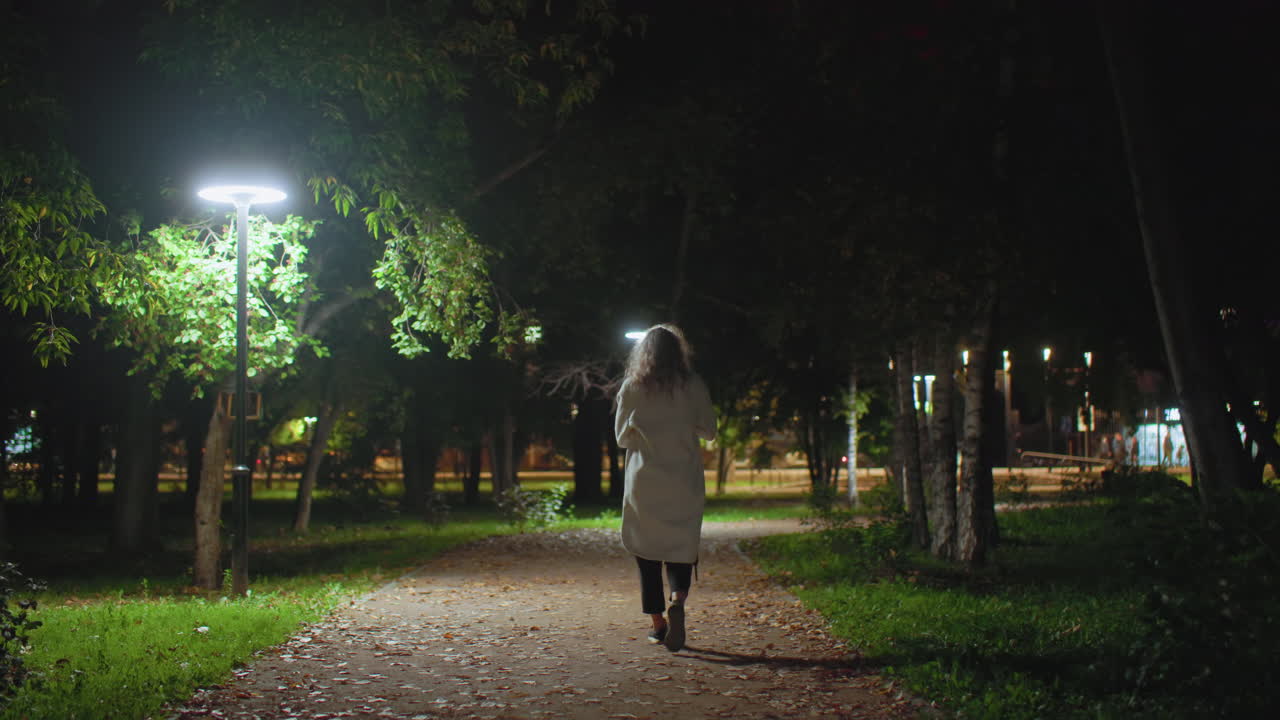 Back view of young adult walking along peaceful paved path under glowing lampstand surrounded by lush green trees at night with blurred motion of car lights in background