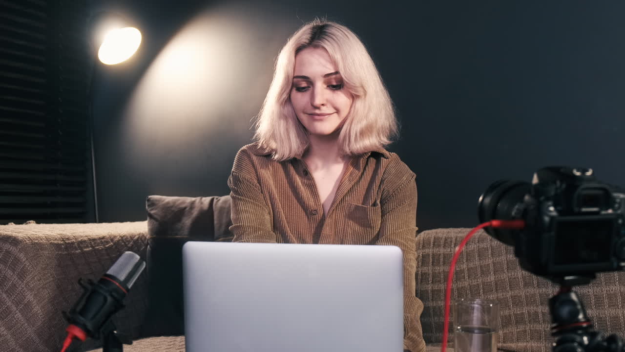 Young content creator blonde girl with cup of water sitting down on the sofa, starting working on her laptop, camera on a tripod, microphone. Working from home