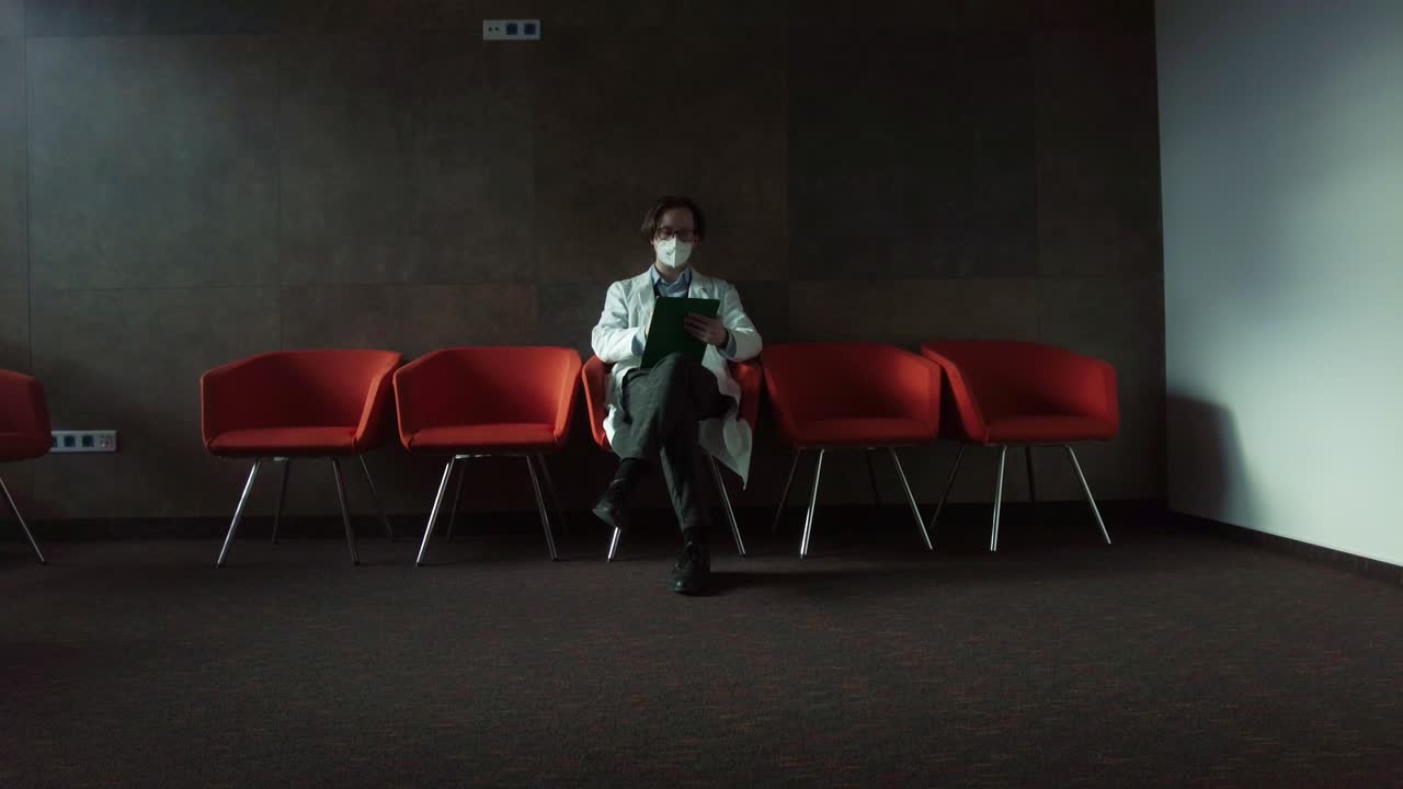 psicoterapeuta con bata blanca y máscara sentada en una silla roja tomando notas en la sala de terapia del hospital