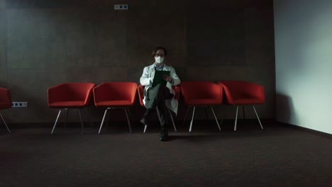 psicoterapeuta con bata blanca y máscara sentada en una silla roja tomando notas en la sala de terapia del hospital