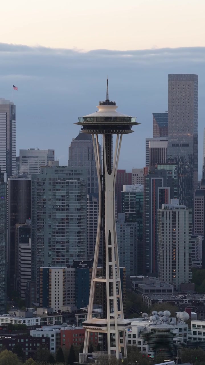 Space Needle seisoo kehystettynä tiheällä Seattlen keskustan pilvenpiirtäjien ryppäällä, lasi- ja terästornien tungeksiessa sen erottuvan lautasenmuotoisen näköalatasanteen alapuolella. Ilmakuva dronella, pystysuuntainen laajakuva.