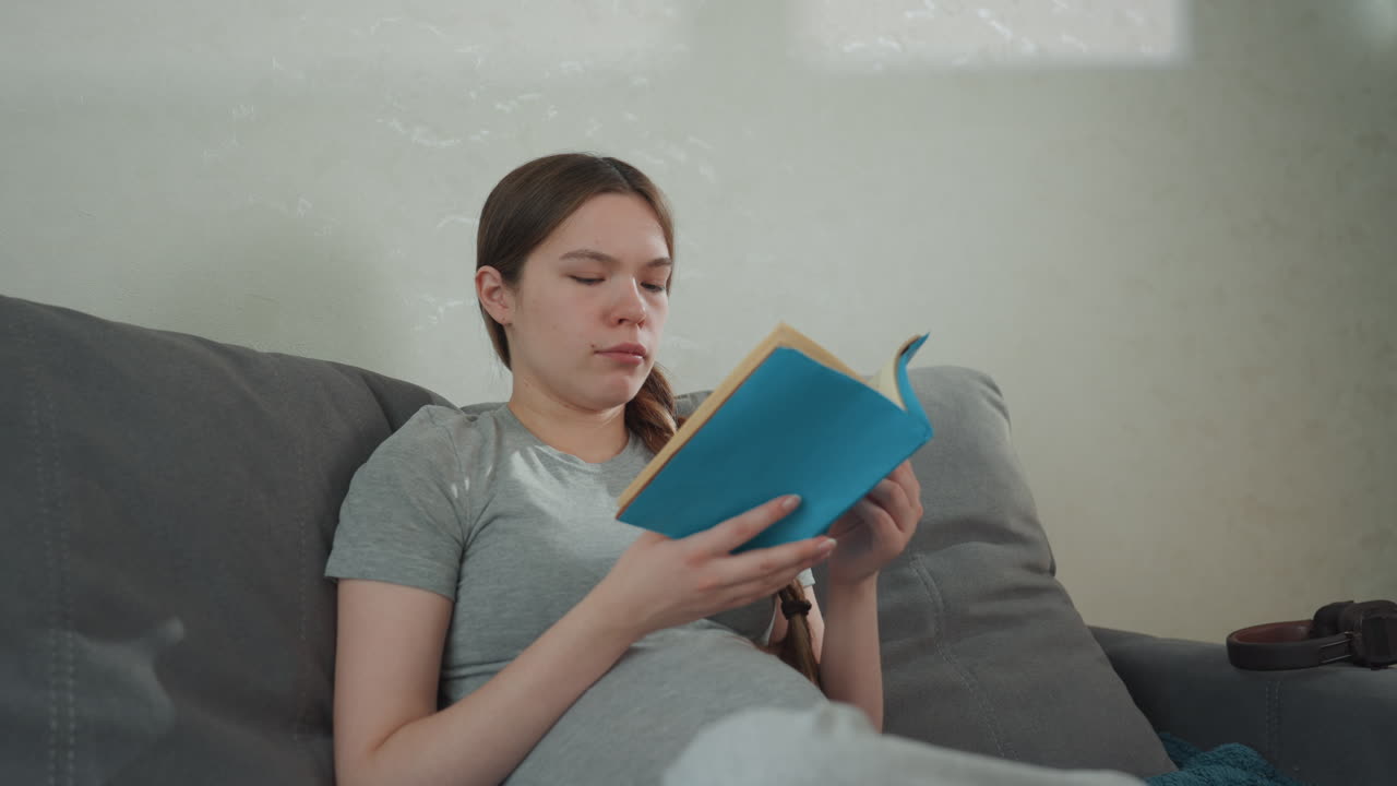 Wanita hamil membaca di sofa, memegang buku biru, postur santai, kaus kasual dan kepang, cahaya sore yang lembut, napas tenang, ketenangan rumah tangga, momen bersarang yang tenang, fokus pada halaman