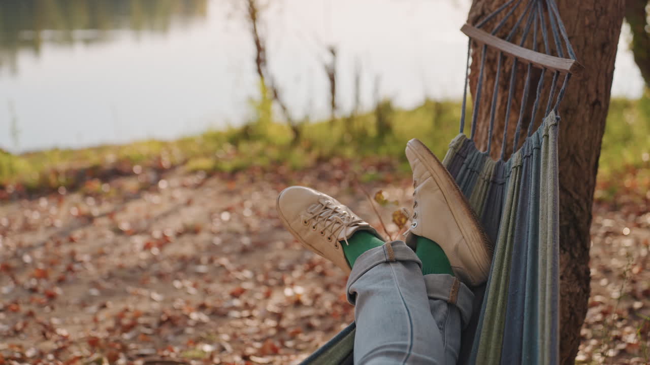 Campeggio con amaca in riva al lago, gambe di campeggiatore rilassato in jeans e scarpe distese, mentre una leggera brezza increspa la corda dell'amaca e l'acqua calma riflette gli alberi, foglie cadute sulla riva, luce solare calda e atmosfera serena
