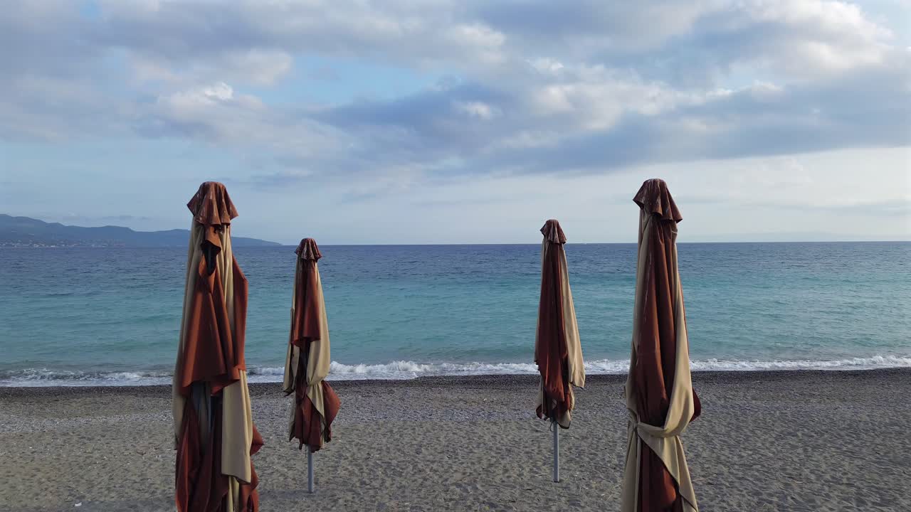 Aerial , low altitude flight between close parasol, beach umbrellas, revealing mesmerizing calm sea at Mesinnian bay, Kalamata, Peloponnesus, Greece. Cloudy day before season summer period 4K