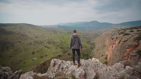 Man-standing-alone-on-cliff-rocks-looking-down-into-valley,-Israel,-aerial-view