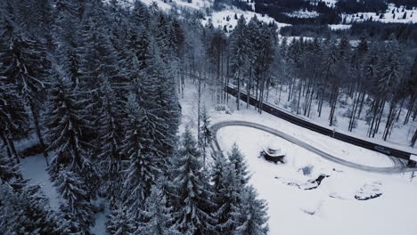 Toma-Aérea-Inclinada-Hacia-Abajo-De-Bosques-Y-Caminos-Nevados-En-Las-Montañas-Bávaras-Durante-La-Temporada-De-Invierno,-Alemania---Automóviles-Circulando-Por-Calles-Con-Curvas