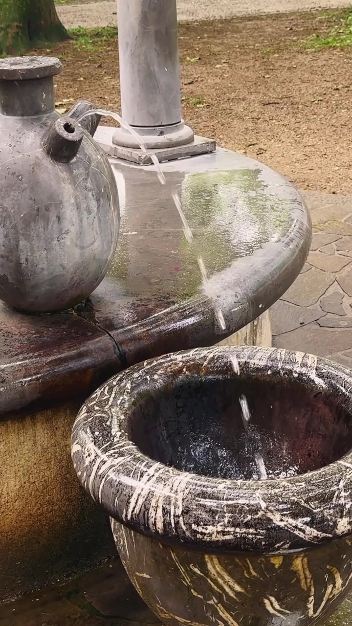 Primo piano verticale di una fontana pubblica ornamentale con acqua che scorre, una vasca in pietra consumata e un elemento scultoreo in un contesto urbano all'aperto