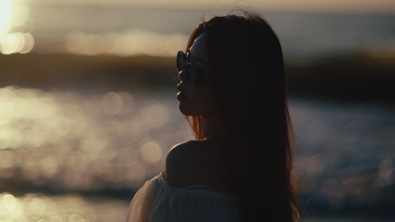 Young woman wearing sunglasses looking toward ocean waves in warm evening light, sideview, beauty model pose with soft smile