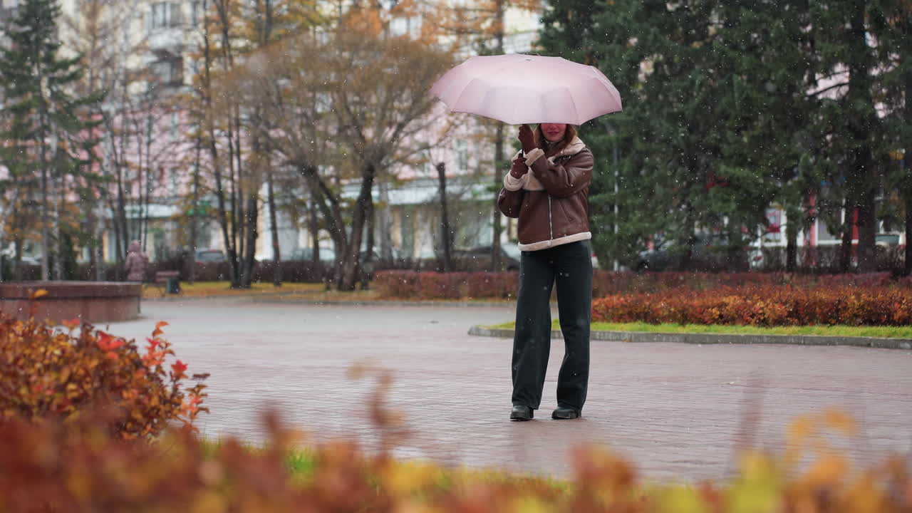 Signora allegra che tiene un ombrello, indossa un berretto di lana, una giacca shearling marrone, pantaloni neri, fa felicemente roteare l'ombrello sotto una leggera nevicata, si gode la giornata invernale, giocosa, diffonde energia positiva nella stagione fredda