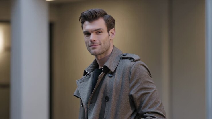 Mann i grå trenchcoat vender seg mot kameraet etter å ha lagt merke til det, smiler med lukket munn i en lobby.