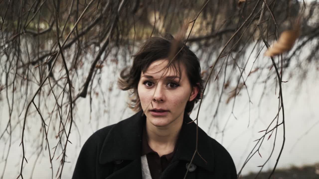 가을 오후 공원의 바람 부는 나무 아래 서 있는 검은 코트를 입은 젊은 여성