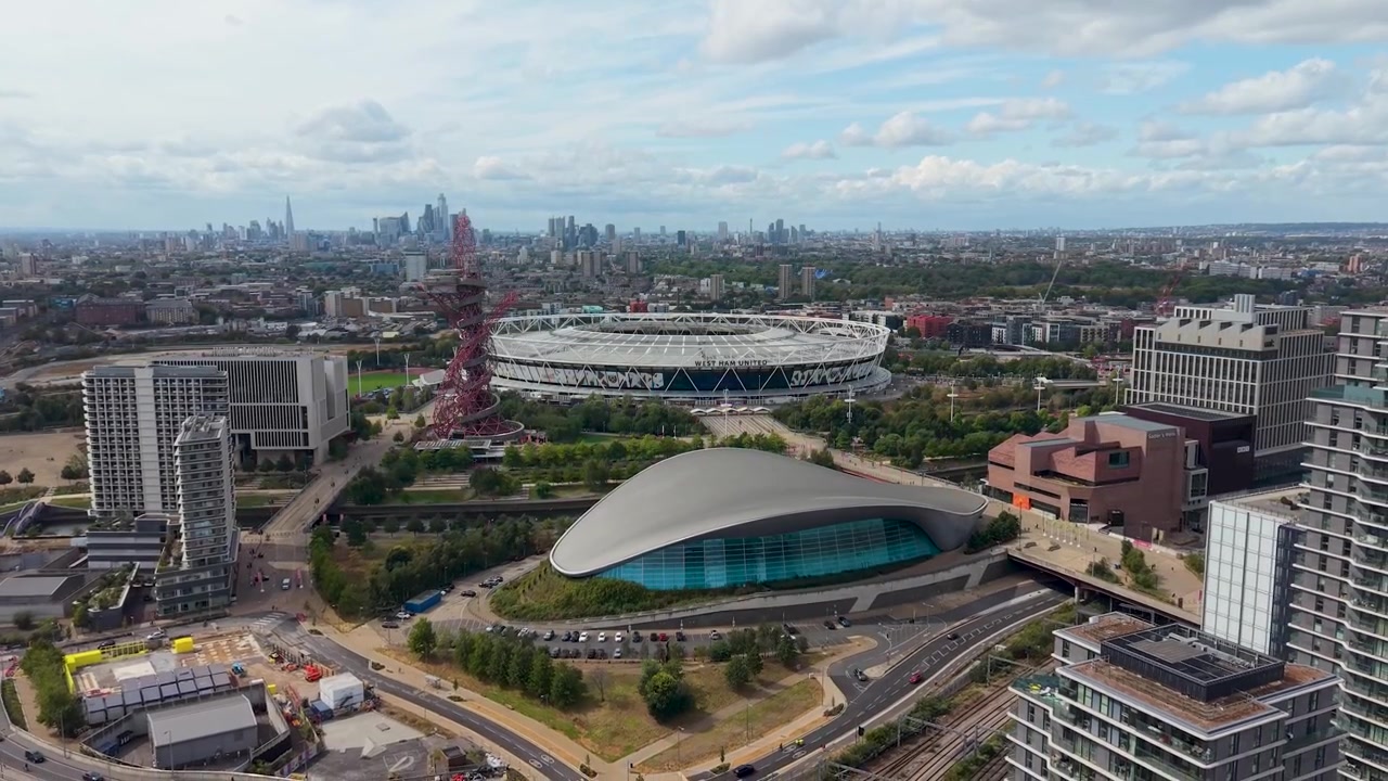 Stadio del West Ham e Centro Acquatico di Londra visti in una ripresa con dolly zoom da drone sopra il Queen Elizabeth Olympic Park, con lo skyline di Londra e strutture moderne sotto nuvole sparse