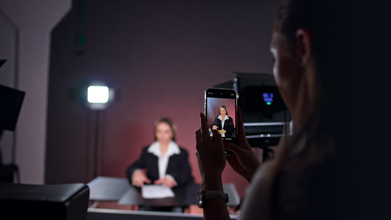 Unrecognized long-haired woman holding a phone and filming a lady at backdrop. Taking pictures of a female reporter working in studio.