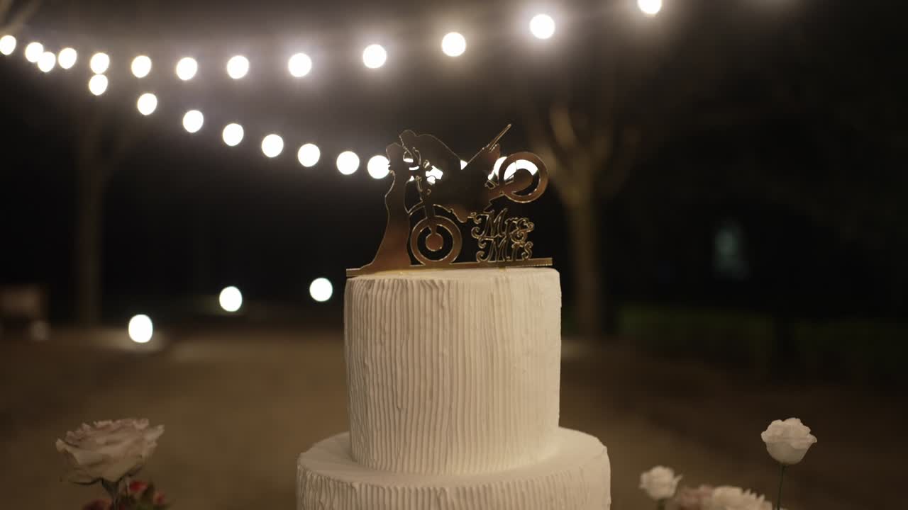 Primo piano di una torta nuziale a due piani con un topper personalizzato, circondata da lucine