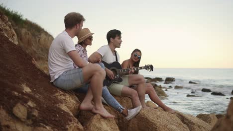 group of young hipster friends sitting on the rocks by the seashore and playing guitar and singing songs. slow motion shot