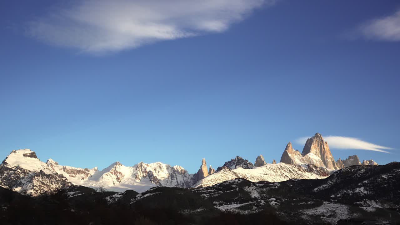 Timelapse dell'iconica catena montuosa Fitz Roy e Cerro Torre in Patagonia, Argentina. Cime innevate illuminate dalla calda luce dell'alba sotto un cielo azzurro e limpido, con un leggero movimento delle nuvole.
