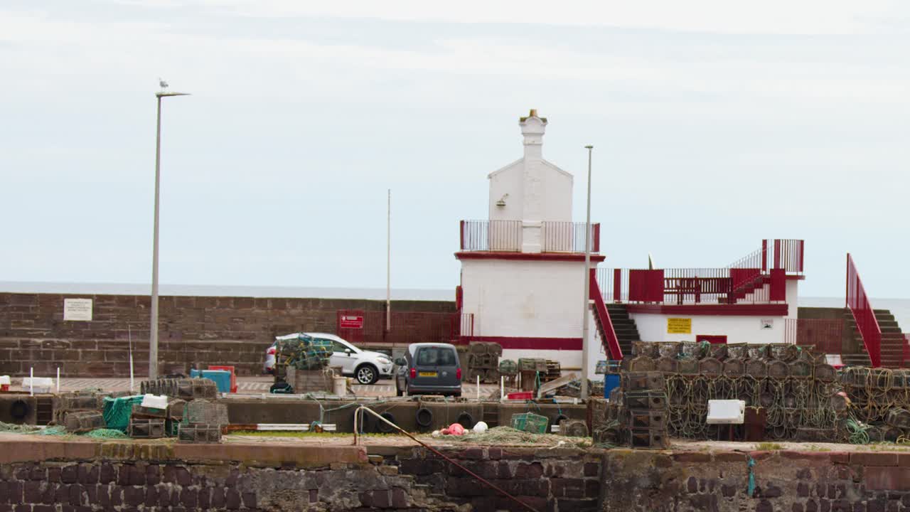 Fishing boat exits harbor past stacked lobster pots, lighthouse, and stone wall under overcast daylight