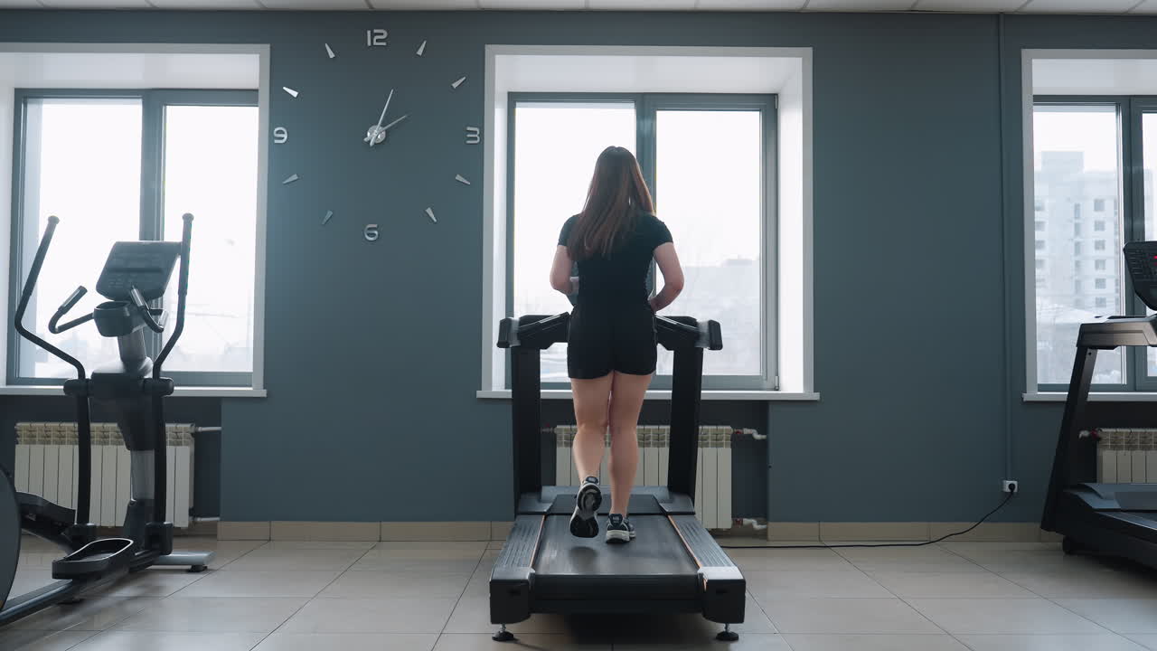 back view of athletic woman exercising on treadmill at high speed in well-lit gym with large wall clock and cardio equipment near windows showing urban buildings in background and minimal interior
