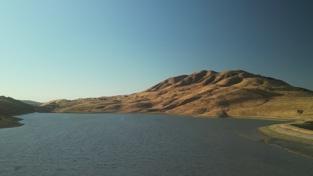 A drone glides over Paicines Reservoir, unveiling its still waters reflecting the sky, surrounded by quiet farmland in the heart of California.