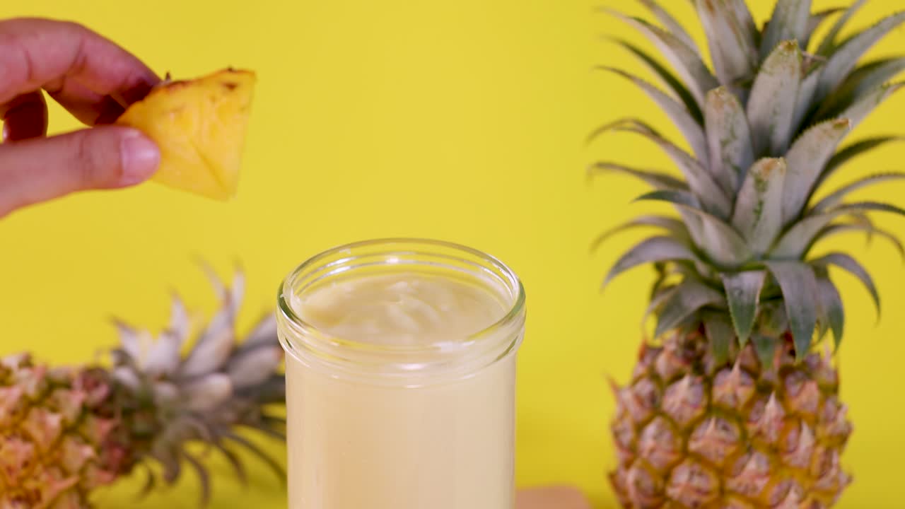 Hand adds pineapple slice to creamy smoothie against vibrant yellow background with fresh pineapples