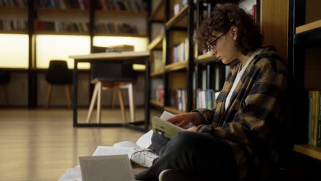 A-focused-brunette-girl-with-curly-hair-wearing-glasses-reads-a-book-in-the-library-among-the-shelves-and-books-on-the-floor