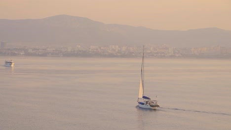 Amanecer,-Puesta-De-Sol-Sobre-El-Adriático-Con-Yate-Flotando-En-El-Mar-Y-Ciudad-Costera-En-El-Fondo