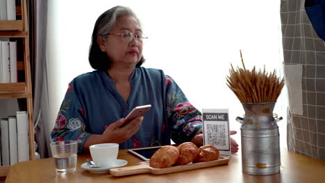 Active-senior-Asian-woman-working-in-a-coffeeshop-using-a-mobile-device,-smartwatch-for-NFC,-QR-code-scanning,-contactless-payment-to-pay-for-coffee-and-snack