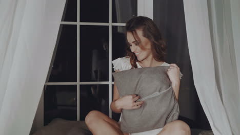 young attractive woman in white nightwear is sitting by the window, posing and smiling in the camera. beautiful woman in lacy pajamas.