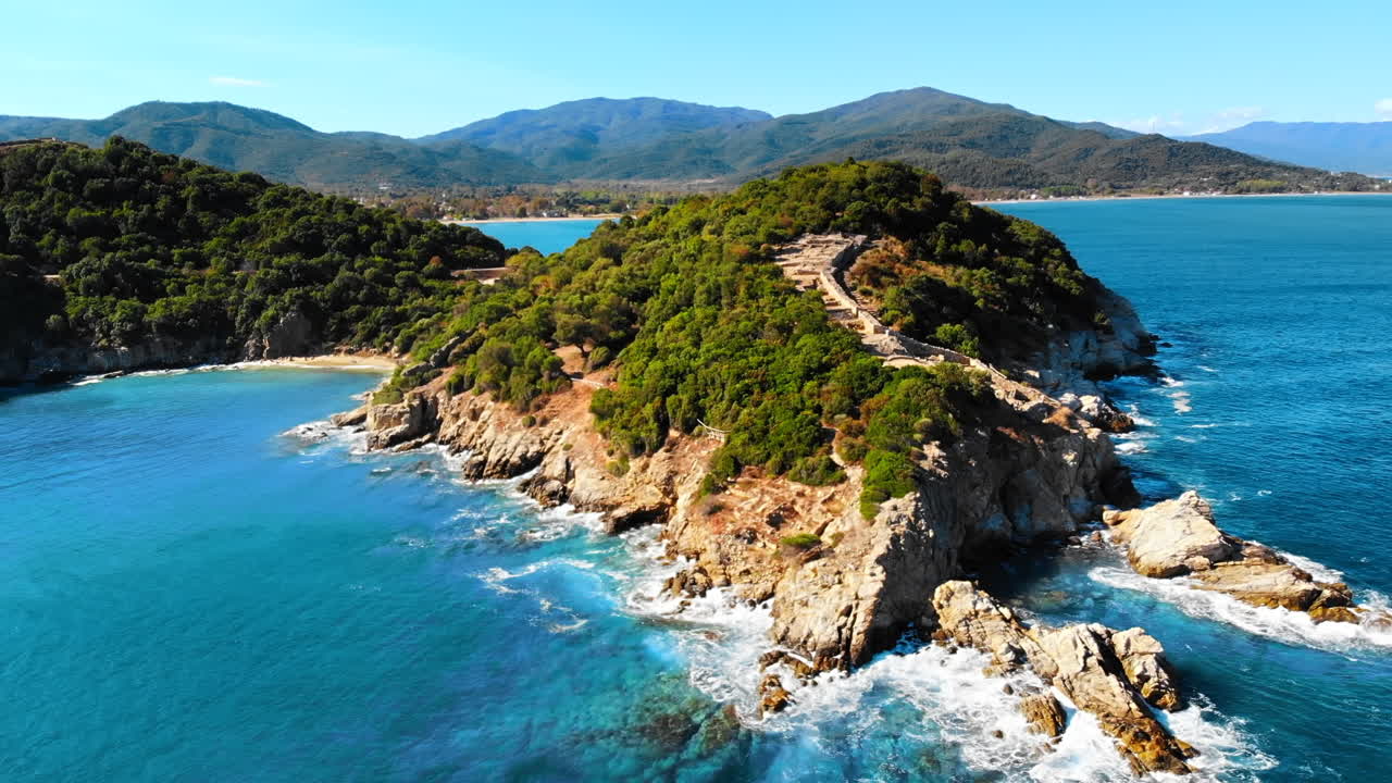 Vista aerea da drone della costa rocciosa del Mar Egeo, penisola con vegetazione e antichi scavi sulla collina, onde con molta schiuma, Grecia