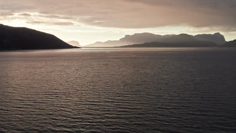 Aerial-shot,-sweeping-over-the-rippling-still-water-of-Sognefjord-in-Norway,-looking-towards-a-sunset-over-shadowy-mountainous-islands-in-the-distance