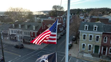 Drone-view-of-an-American-and-POW-flag-raised-at-half-staff-above-this-American-small-town-surrounded-by-a-close-knit-community-at-sunset