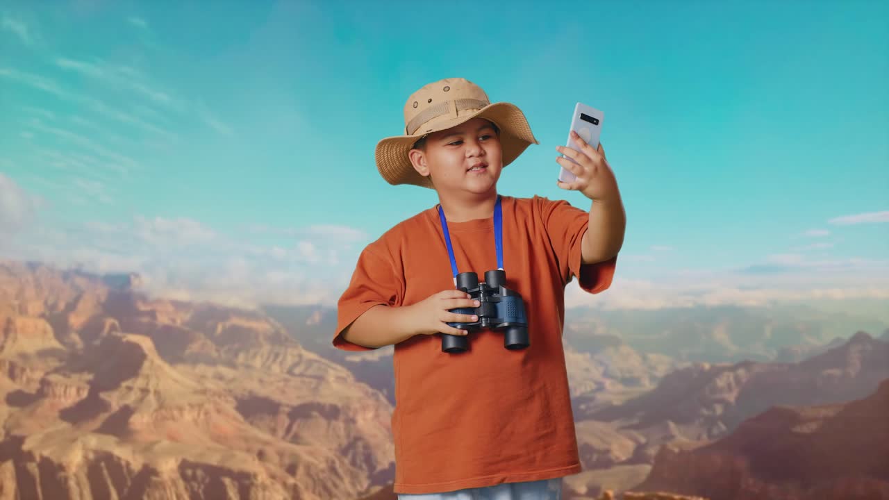 Asian Boy With A Hat And Binoculars Waving Hand Having Video Call On Smartphone While Traveling At The Top Of Mountain. Boy Researcher Examines Something, Travel Tourism Adventure