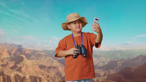 asian boy with a hat and binoculars waving hand having video call on smartphone while traveling at the top of mountain. boy researcher examines something, travel tourism adventure