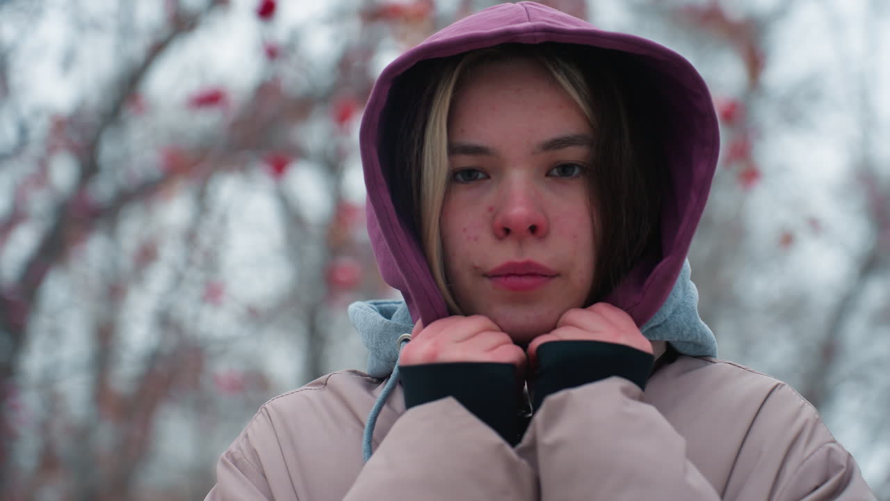 냉정한 표정으로 겨울 재을 입고 야외에 서있는 젊은 여자의 가까운 사진, 후디 목걸이를 들고, 카메라를 직접 바라보고, 배경에 흐릿한 나무, 추운 날씨, 부드러운 가을 분위기