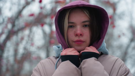 primer plano de una joven de pie al aire libre con chaqueta de invierno con mirada sobria, sosteniendo el cuello de la capucha, mirando directamente a la cámara, árboles borrosos en el fondo, clima frío, suave atmósfera de otoño