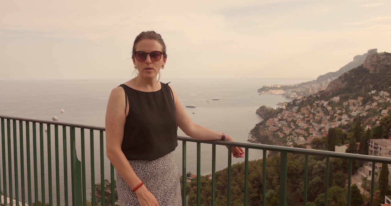 Woman informative talks to camera at a hilltop railing with the French Riviera and Monaco coastline stretching out behind her overlooking the French Riviera, Roquebrune-Cap-Martin, France