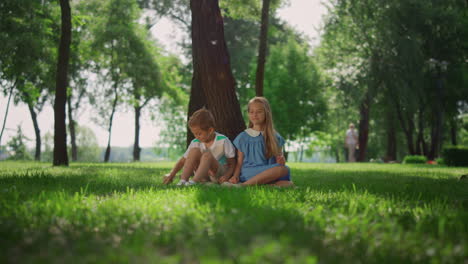 Lachende-Kinder-Sitzen-Unter-Einem-Baum-Im-Grünen-Park.-Glückliches,-Aktives-Kindheitskonzept.