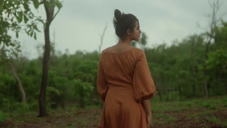 A-woman-in-a-brown-dress-stands-in-a-lush-green-forest-during-a-calm-day