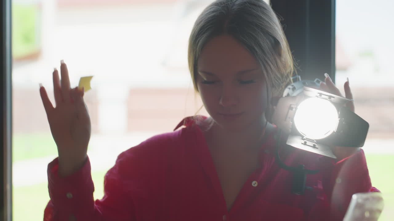 Young woman in red shirt holds bright spotlight over shoulder while eating sliced cheese near large window, natural light blends with artificial beam
