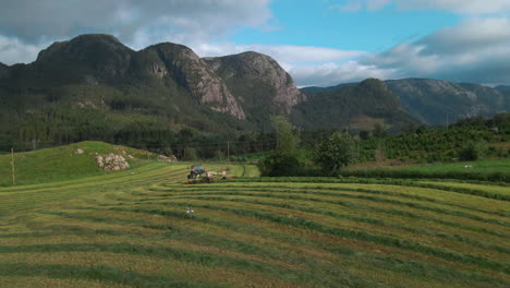 Grass-turner-raking-grass-for-silage-production-on-scenic-farm,-aerial-slowmo