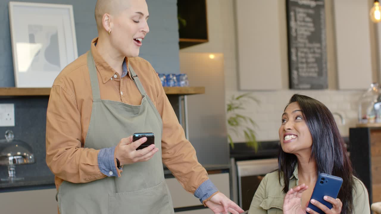 Indian woman scrolling phone at cafe with croissant to-go cup, woman barista leaning showing screen