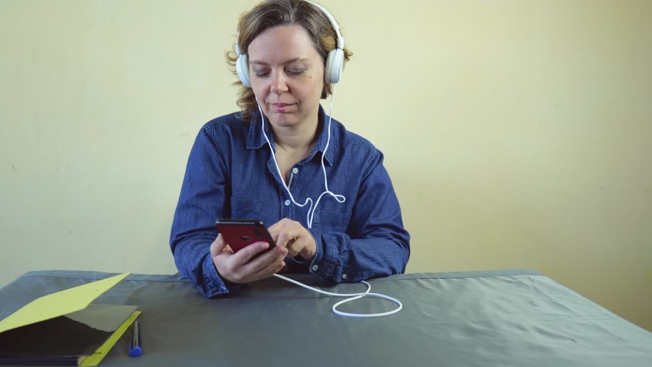 A forty-year-old woman in wired large white headphones is sitting at a table listening to music. An adult woman is holding a mobile phone
