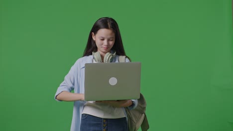 estudiante adolescente asiática con una mochila sonriendo y escribiendo en una computadora portátil mientras está de pie en el estudio de fondo de la pantalla verde