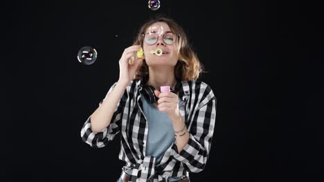 retrato de una chica feliz con gafas soplando muchas burbujas de jabón con un palo en fondo negro. imágenes de estudio de una morena con cabello corto y labios sensuales con auriculares en el cuello. cámara lenta