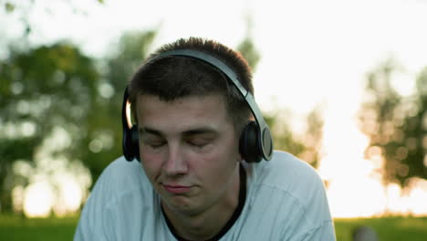 close up of music artist lying on grass wearing headphones nodding to music while using phone, looking upward thoughtfully, with soft bokeh effect from trees