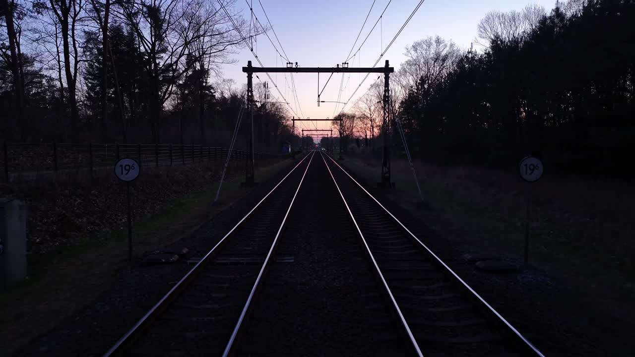 View from a train at dusk crossing a road, surrounded by trees and railway tracks in Ochten.