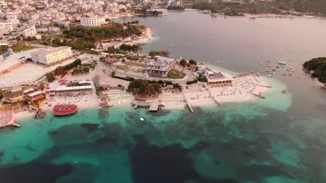 Drone-view-in-Albania-flying-over-blue-crystal-clear-water-on-sunset,-green-hills,-boats-and-hotels-in-Ksamil